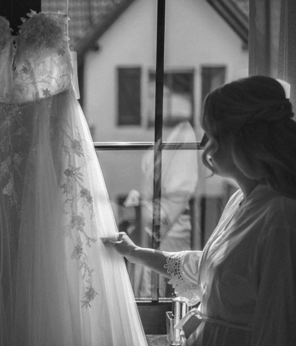 Braut steht vor ihrem Hochzeitskleid, welches am Fenster hängt